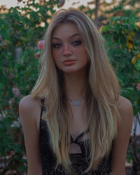 a girl with long blonde hair standing in front of bushes